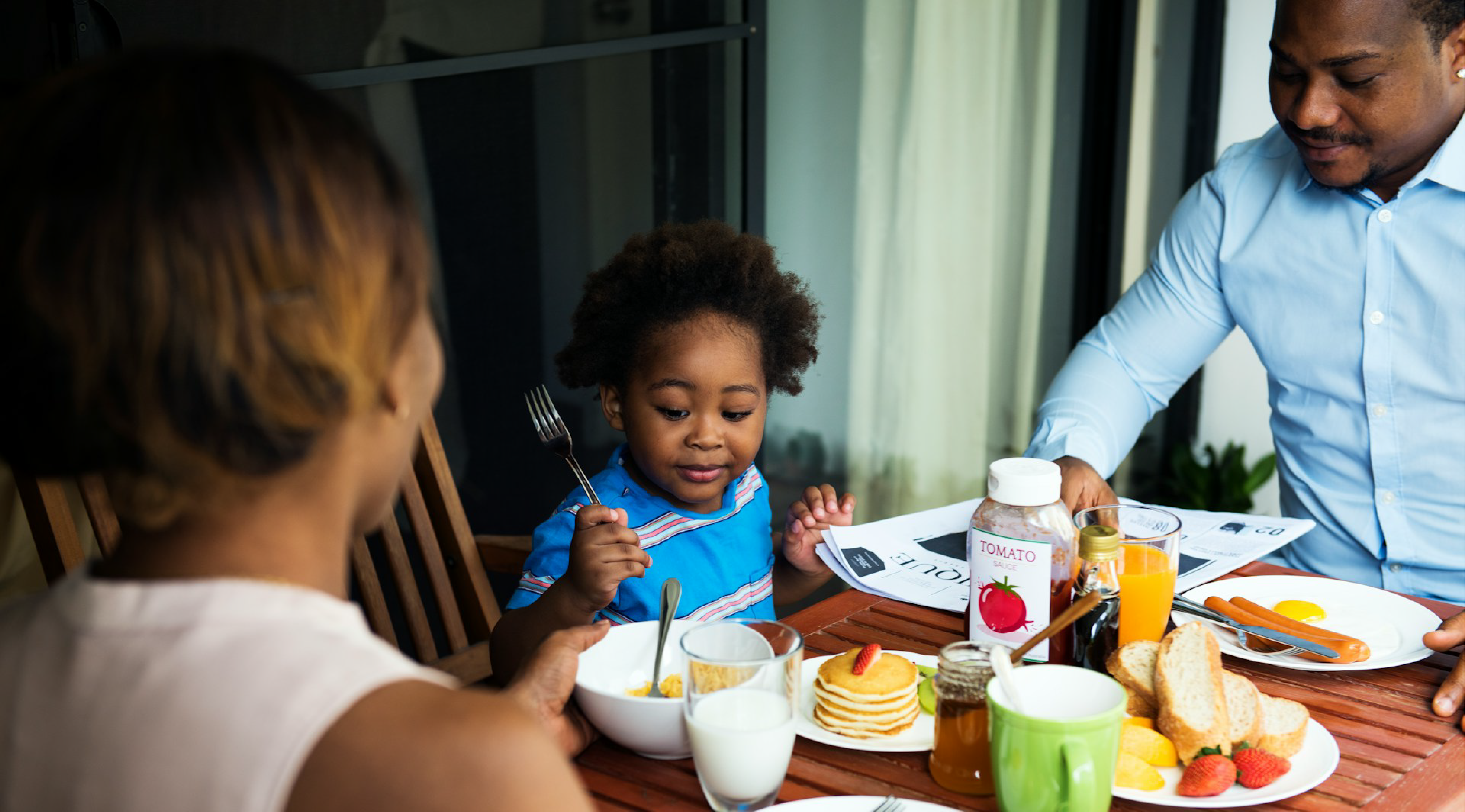 An African American family sits around a breakfast table; the woman’s back faces the viewer, a child in the middle holds up a fork while eating, and the man on the right reads a newspaper. The table is set with pancakes, orange juice, and other breakfast foods.