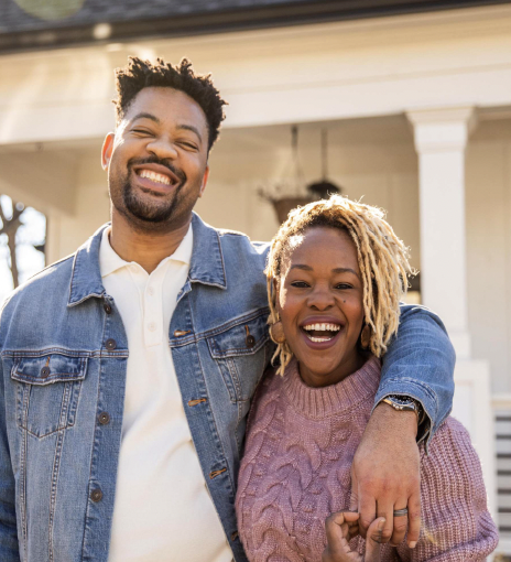 A joyful African American couple stands in front of their first home; the man wraps his arm around the woman as she holds his hands, both smiling with excitement and pride.