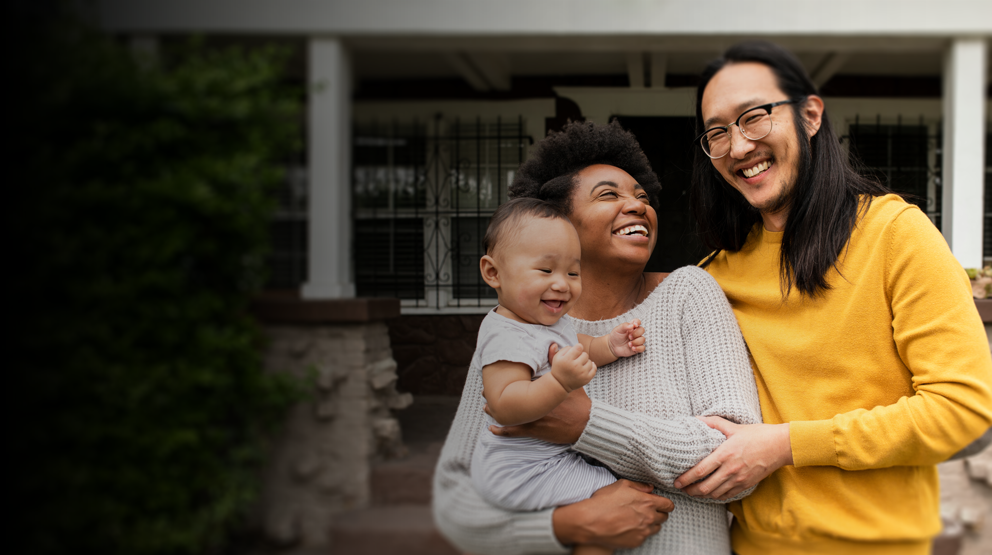 A joyful young family stands in front of their newly purchased home; the father embraces the mother, who is holding their smiling baby, all radiating happiness and excitement.